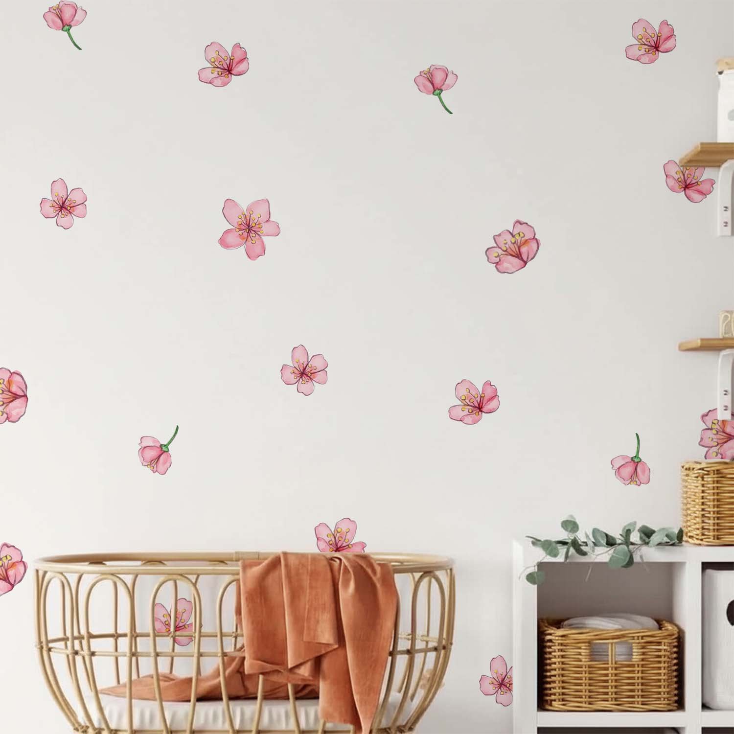 Scattered pink cherry blossom wall stickers decorating a soft pastel nursery wall beside a wicker chair, knitted blanket, and pink canopy crib.