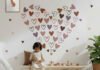 A children's bedroom featuring pink, brown, and mauve heart wall stickers arranged in a large heart shape above a wooden bench, creating a cozy boho nursery decor theme.