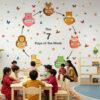 Colorful educational wall sticker featuring the 7 days of the week arranged in a circular sequence with cute animal characters, butterflies, bees, and flowers displayed above a wooden cabinet in a children's playroom with a mother and three children.