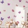 Purple and pink watercolor butterfly wall stickers arranged on a white bedroom wall above a bed, featuring removable peel-and-stick vinyl butterfly decals for girls' rooms, nurseries, and home décor.