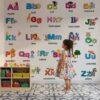 Colorful A–Z alphabet educational wall sticker with illustrated objects for each letter displayed in a preschool classroom where a teacher is teaching children using peel-and-stick learning wall decals.