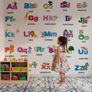 Colorful A–Z alphabet educational wall sticker with illustrated objects for each letter displayed in a preschool classroom where a teacher is teaching children using peel-and-stick learning wall decals.